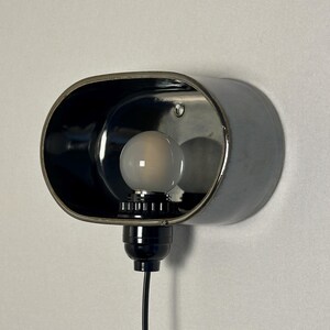 K&ouml;nnte beinhalten: Eine wandmontierte, ovale Lampe mit schwarzem Metallfinish. Die Lampe hat eine Gl&uuml;hbirne und ein schwarzes Kabel. Das Innere der Lampe ist reflektierend und an einer wei&szlig;en Wand befestigt.