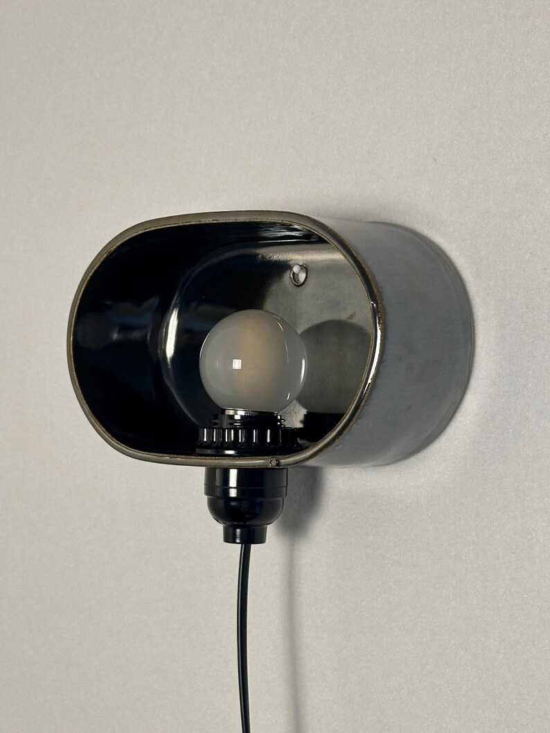 K&ouml;nnte beinhalten: Eine wandmontierte, ovale Lampe mit schwarzem Metallfinish. Die Lampe hat eine Gl&uuml;hbirne und ein schwarzes Kabel. Das Innere der Lampe ist reflektierend und an einer wei&szlig;en Wand befestigt.