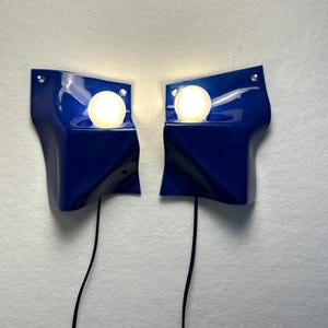 May include: A pair of wall-mounted lamps featuring glossy, deep blue, wave-shaped ceramic shades. Each lamp includes a single, spherical, glowing white light bulb positioned within a recessed circular opening. Thin black power cords extend downward.