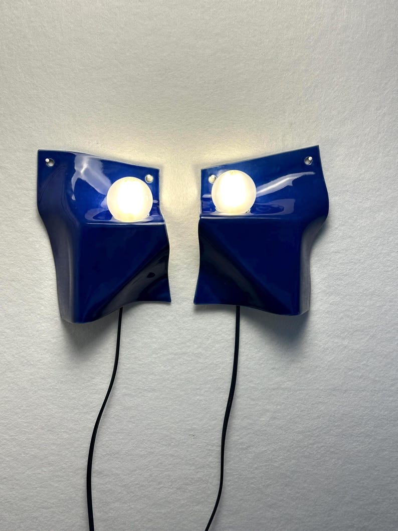 May include: A pair of wall-mounted lamps featuring glossy, deep blue, wave-shaped ceramic shades. Each lamp includes a single, spherical, glowing white light bulb positioned within a recessed circular opening. Thin black power cords extend downward.