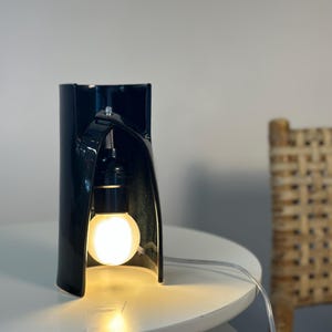May include: A glossy black ceramic table lamp with a curved, cylindrical structure that features an arched cutout. A single round, glowing light bulb is suspended within the opening, connected by a clear electrical cord extending to the side.