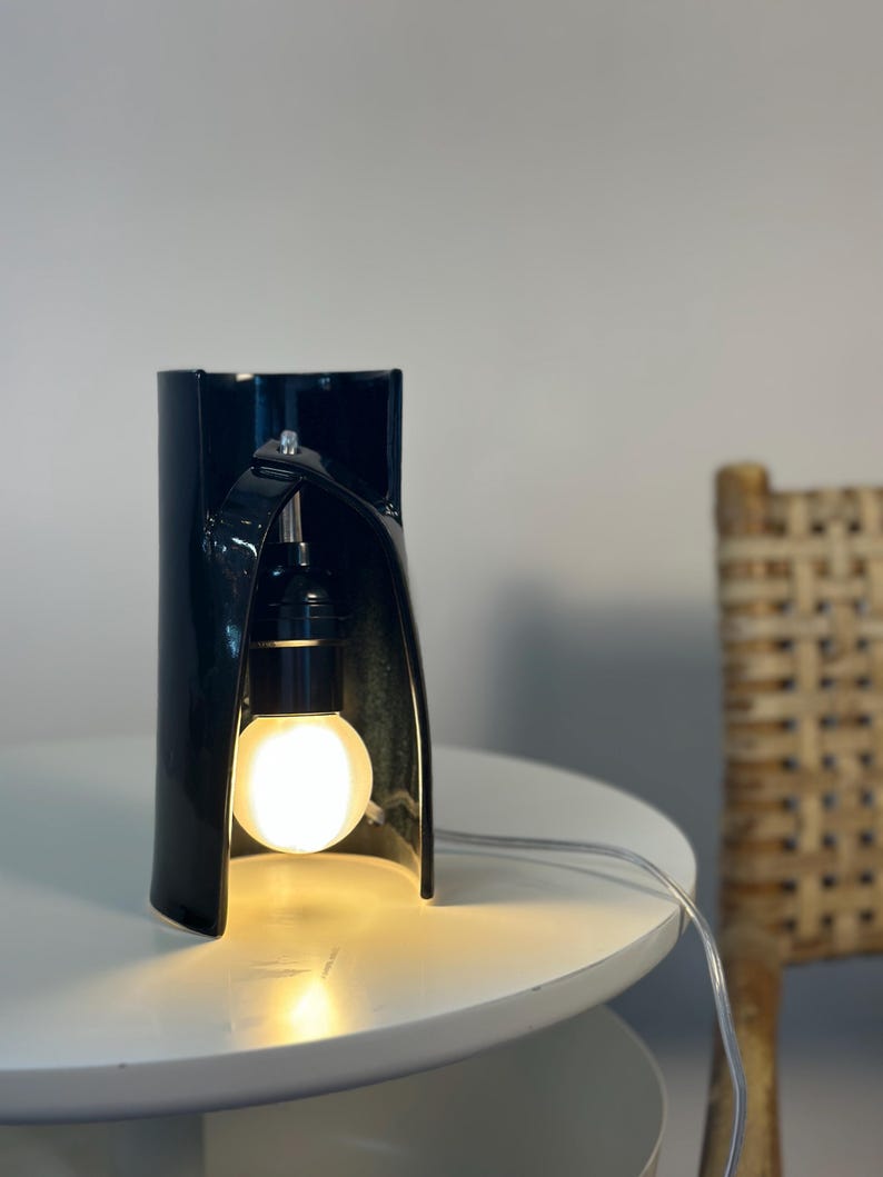 May include: A glossy black ceramic table lamp with a curved, cylindrical structure that features an arched cutout. A single round, glowing light bulb is suspended within the opening, connected by a clear electrical cord extending to the side.