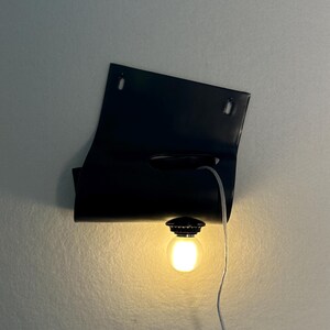 Puede incluir: Una l&aacute;mpara de pared negra con una pantalla de metal rectangular y curva que tiene dos orificios de montaje en la parte superior. Una bombilla redonda e iluminada est&aacute; sujeta al borde inferior y un cable fino y transparente cuelga hacia abajo.