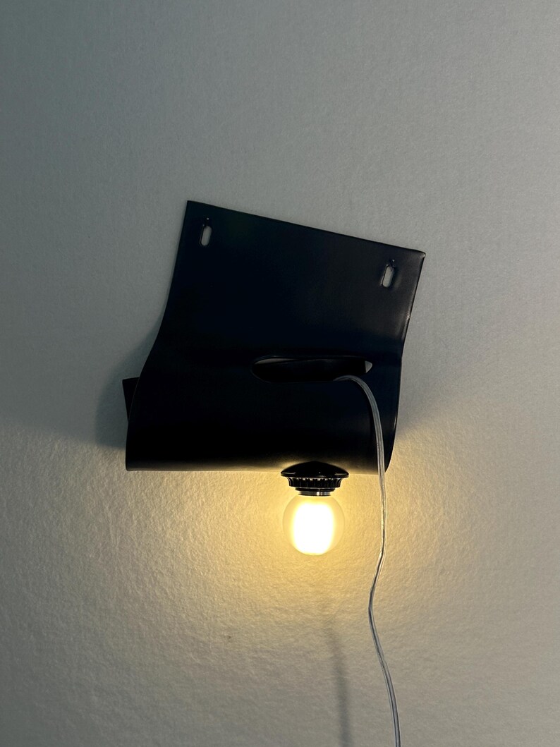 Puede incluir: Una l&aacute;mpara de pared negra con una pantalla de metal rectangular y curva que tiene dos orificios de montaje en la parte superior. Una bombilla redonda e iluminada est&aacute; sujeta al borde inferior y un cable fino y transparente cuelga hacia abajo.
