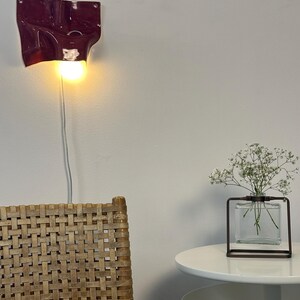 May include: A deep burgundy wall-mounted ceramic lamp with a fluid, organic shape and a single glowing light bulb. The fixture features a smooth, glossy finish and is connected to a thin white power cord that hangs vertically down the wall.