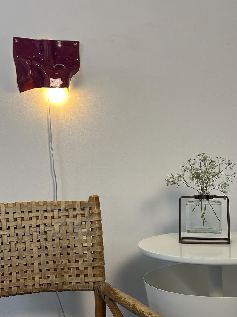 May include: A deep burgundy wall-mounted ceramic lamp with a fluid, organic shape and a single glowing light bulb. The fixture features a smooth, glossy finish and is connected to a thin white power cord that hangs vertically down the wall.