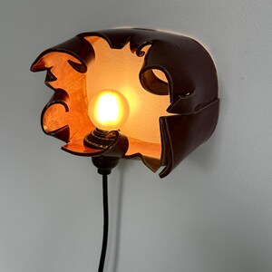 May include: A unique, handcrafted wall sconce with a dark brown, petal-like shade. The interior glows with warm orange light, housing a round, illuminated bulb. A black cord extends downwards, completing the design. This decorative lighting fixture adds a touch of artistry.