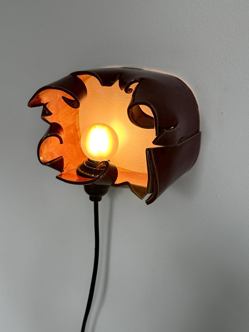 May include: A unique, handcrafted wall sconce with a dark brown, petal-like shade. The interior glows with warm orange light, housing a round, illuminated bulb. A black cord extends downwards, completing the design. This decorative lighting fixture adds a touch of artistry.