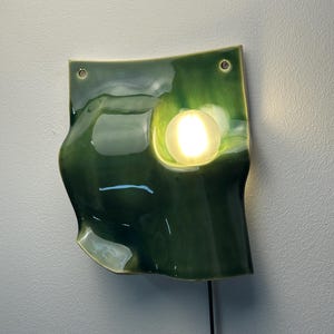 May include: A wall-mounted ceramic light fixture with a glossy, deep green glaze and an undulating, organic shape. The piece features two circular holes at the top for mounting and a single, round, illuminated light bulb set into a recessed area.