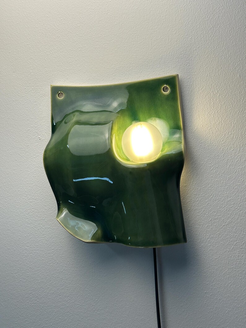 May include: A wall-mounted ceramic light fixture with a glossy, deep green glaze and an undulating, organic shape. The piece features two circular holes at the top for mounting and a single, round, illuminated light bulb set into a recessed area.