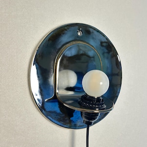 May include: A circular wall sconce featuring a glossy, deep blue and black glazed ceramic backplate with a small hole at the top. A small, round, frosted white light bulb is mounted on a black socket attached to a protruding ceramic shelf.