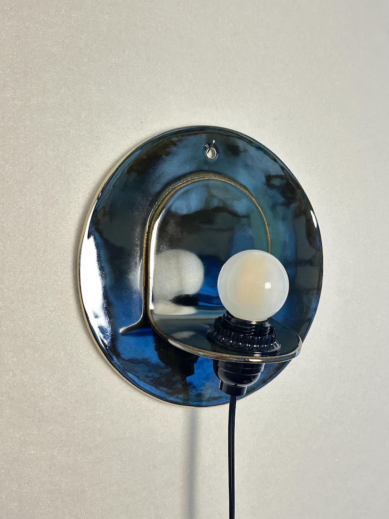 May include: A circular wall sconce featuring a glossy, deep blue and black glazed ceramic backplate with a small hole at the top. A small, round, frosted white light bulb is mounted on a black socket attached to a protruding ceramic shelf.