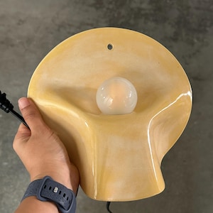 May include: A wall-mounted lamp featuring a smooth, pale yellow, shell-shaped ceramic shade with a central circular opening for a small, round, frosted white light bulb. The fixture includes a black electrical cord and a small hole for mounting.