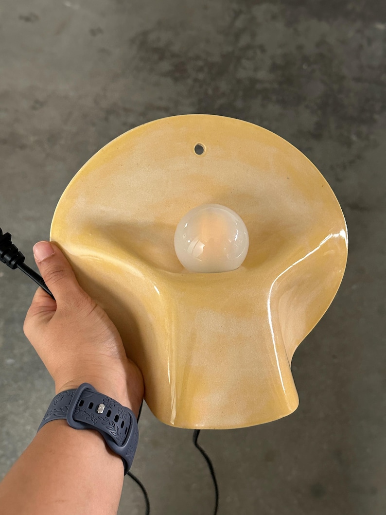 May include: A wall-mounted lamp featuring a smooth, pale yellow, shell-shaped ceramic shade with a central circular opening for a small, round, frosted white light bulb. The fixture includes a black electrical cord and a small hole for mounting.