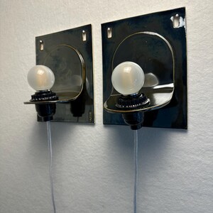 May include: Two wall-mounted sconces with a dark blue finish. Each sconce features a curved, metallic shade, a black bulb socket, and a frosted glass bulb. A silver wire extends from each fixture. The sconces are mounted on a white wall.