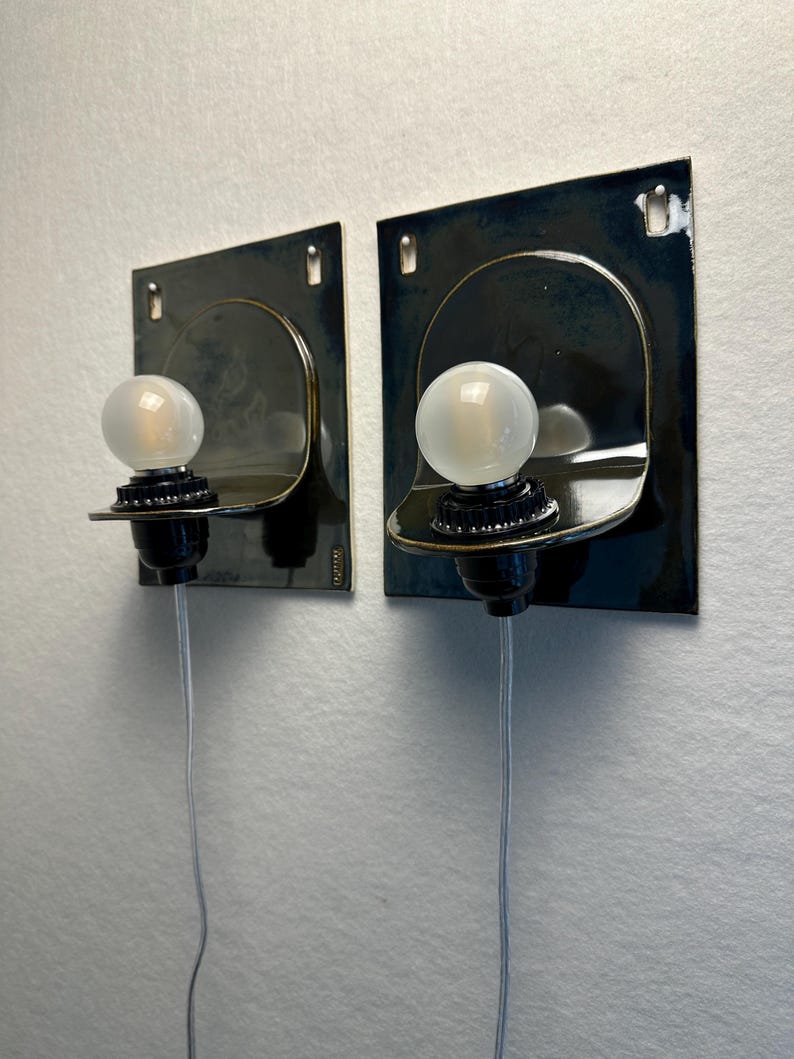 May include: Two wall-mounted sconces with a dark blue finish. Each sconce features a curved, metallic shade, a black bulb socket, and a frosted glass bulb. A silver wire extends from each fixture. The sconces are mounted on a white wall.