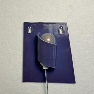 Pode incluir: Uma arandela de parede moderna com uma placa traseira retangular roxa escura e dois orif&iacute;cios de montagem. Uma sombra curva roxa abriga uma l&acirc;mpada esf&eacute;rica fosca. Um fio fino e prateado se estende da base.