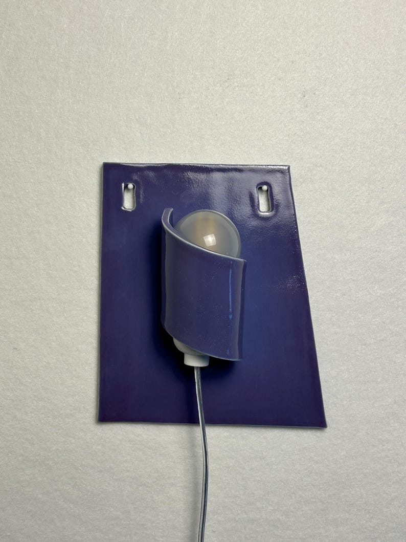 Pode incluir: Uma arandela de parede moderna com uma placa traseira retangular roxa escura e dois orif&iacute;cios de montagem. Uma sombra curva roxa abriga uma l&acirc;mpada esf&eacute;rica fosca. Um fio fino e prateado se estende da base.