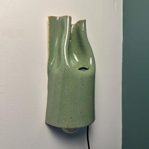 May include: A wall-mounted light fixture made of light green ceramic with a speckled finish. The fixture has a vertical, undulating, folded shape with a smooth, glossy surface. A single light bulb is visible at the bottom with a black power cord.