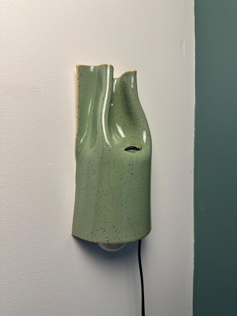 May include: A wall-mounted light fixture made of light green ceramic with a speckled finish. The fixture has a vertical, undulating, folded shape with a smooth, glossy surface. A single light bulb is visible at the bottom with a black power cord.