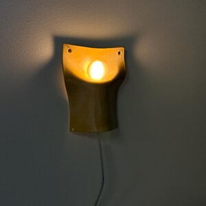 May include: A mustard yellow ceramic wall sconce with a curved, organic shape and a central circular cutout. A warm, glowing light bulb is positioned within the center of the fixture, casting a soft illumination and shadow against the wall.