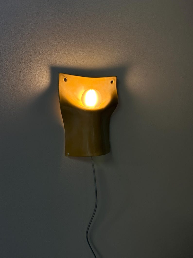 May include: A mustard yellow ceramic wall sconce with a curved, organic shape and a central circular cutout. A warm, glowing light bulb is positioned within the center of the fixture, casting a soft illumination and shadow against the wall.