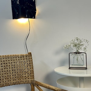 Puede incluir: Un aplique de pared negro con forma ondulada y escultural que emite una luz c&aacute;lida, situado sobre una silla de rat&aacute;n trenzado. A la derecha, un jarr&oacute;n cuadrado de cristal transparente con marco met&aacute;lico contiene flores blancas sobre una mesa.
