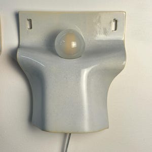 May include: A wall-mounted ceramic light fixture with a glossy, pale blue-grey finish. The piece features a curved, sculptural form with two rectangular mounting holes at the top and a central circular opening holding a frosted glass light bulb.