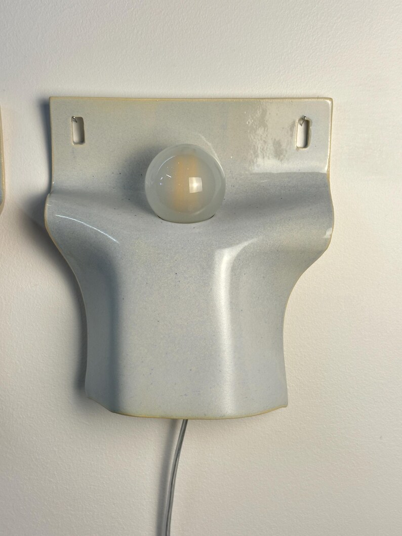 May include: A wall-mounted ceramic light fixture with a glossy, pale blue-grey finish. The piece features a curved, sculptural form with two rectangular mounting holes at the top and a central circular opening holding a frosted glass light bulb.