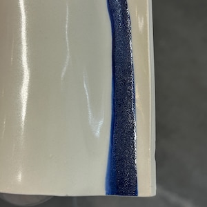 May include: A smooth, off-white ceramic surface featuring a vertical, textured stripe of deep blue glaze. The stripe has a granular, matte appearance, contrasting with the glossy, reflective finish of the surrounding cream-colored ceramic material.