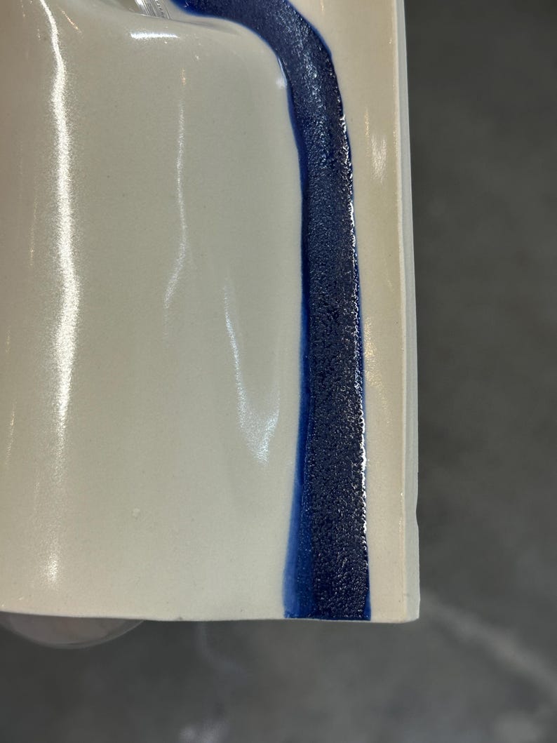 May include: A smooth, off-white ceramic surface featuring a vertical, textured stripe of deep blue glaze. The stripe has a granular, matte appearance, contrasting with the glossy, reflective finish of the surrounding cream-colored ceramic material.