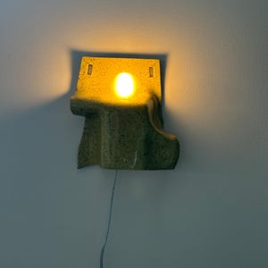 Puede incluir: Un aplique de pared de cer&aacute;mica con un esmalte moteado en amarillo y verde. La l&aacute;mpara presenta una forma org&aacute;nica irregular con una abertura circular central que emite una luz c&aacute;lida. Un cable fino cuelga del borde inferior.