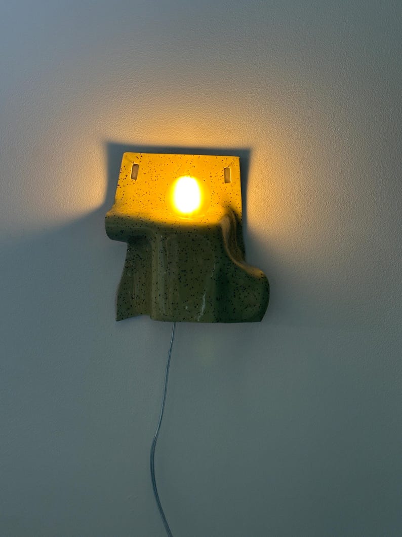 Puede incluir: Un aplique de pared de cer&aacute;mica con un esmalte moteado en amarillo y verde. La l&aacute;mpara presenta una forma org&aacute;nica irregular con una abertura circular central que emite una luz c&aacute;lida. Un cable fino cuelga del borde inferior.