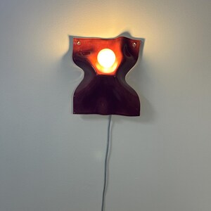 Puede incluir: Un aplique de pared de color rojo oscuro con bordes ondulados y un recorte circular central que emite una luz c&aacute;lida y brillante desde una bombilla interna. El accesorio est&aacute; montado en la pared e incluye un cable fino de color claro.