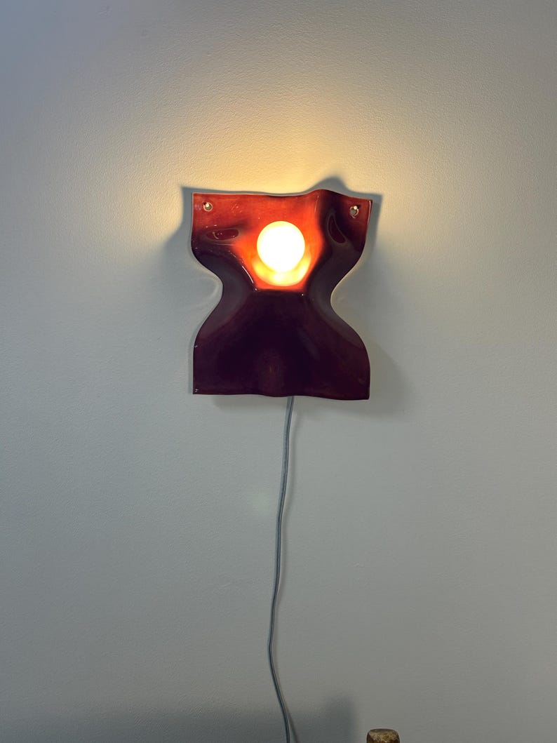 Puede incluir: Un aplique de pared de color rojo oscuro con bordes ondulados y un recorte circular central que emite una luz c&aacute;lida y brillante desde una bombilla interna. El accesorio est&aacute; montado en la pared e incluye un cable fino de color claro.
