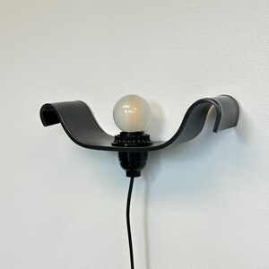 May include: Black wall-mounted light fixture with a curved, wave-like design. The fixture holds a round, frosted light bulb. A black electrical cord hangs down from the fixture against a white wall. The fixture is a modern design.
