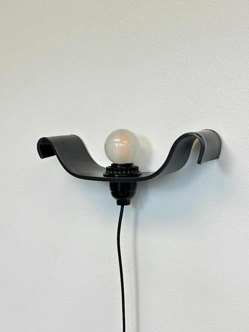 May include: Black wall-mounted light fixture with a curved, wave-like design. The fixture holds a round, frosted light bulb. A black electrical cord hangs down from the fixture against a white wall. The fixture is a modern design.