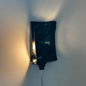 May include: A wall-mounted sconce featuring a glossy, dark-colored, curved ceramic shade. The fixture emits a warm, glowing light from within, casting soft illumination upward and downward against the wall, with a thin power cord extending below.