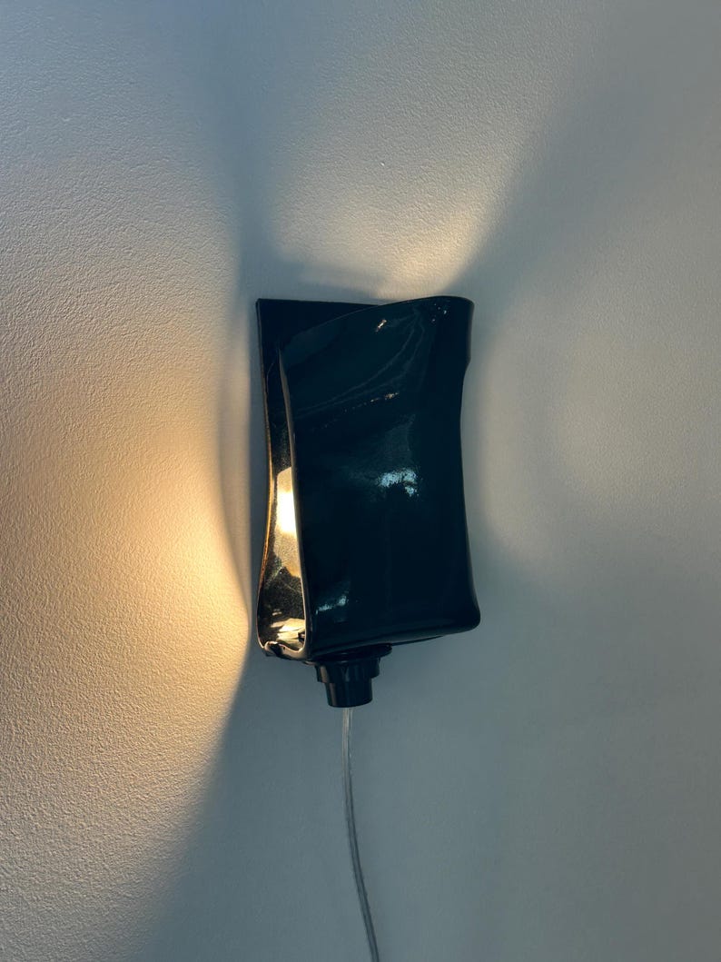 May include: A wall-mounted sconce featuring a glossy, dark-colored, curved ceramic shade. The fixture emits a warm, glowing light from within, casting soft illumination upward and downward against the wall, with a thin power cord extending below.