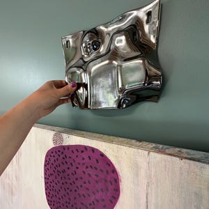Pu&ograve; includere: Un oggetto da parete metallico, riflettente e color argento con una superficie irregolare, ondulata e deformata. Il pezzo presenta due aperture rettangolari vicino al bordo superiore per il fissaggio e mostra una finitura a specchio distorta.