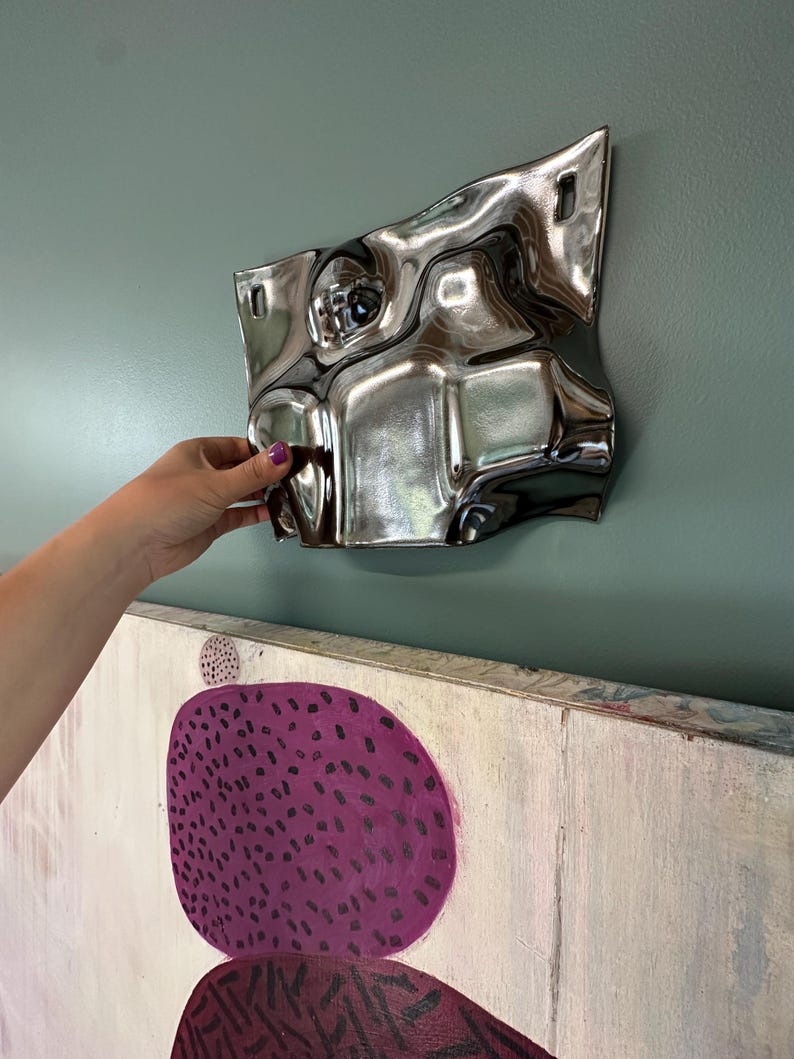 Pu&ograve; includere: Un oggetto da parete metallico, riflettente e color argento con una superficie irregolare, ondulata e deformata. Il pezzo presenta due aperture rettangolari vicino al bordo superiore per il fissaggio e mostra una finitura a specchio distorta.