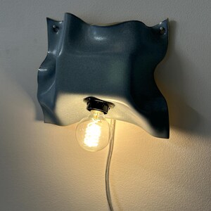 May include: A wall-mounted lamp featuring a blue, wavy, ceramic-like shade with a glossy finish. The fixture includes a black socket holding a glowing, round, clear glass light bulb. A white electrical cord hangs down from the base of the light.