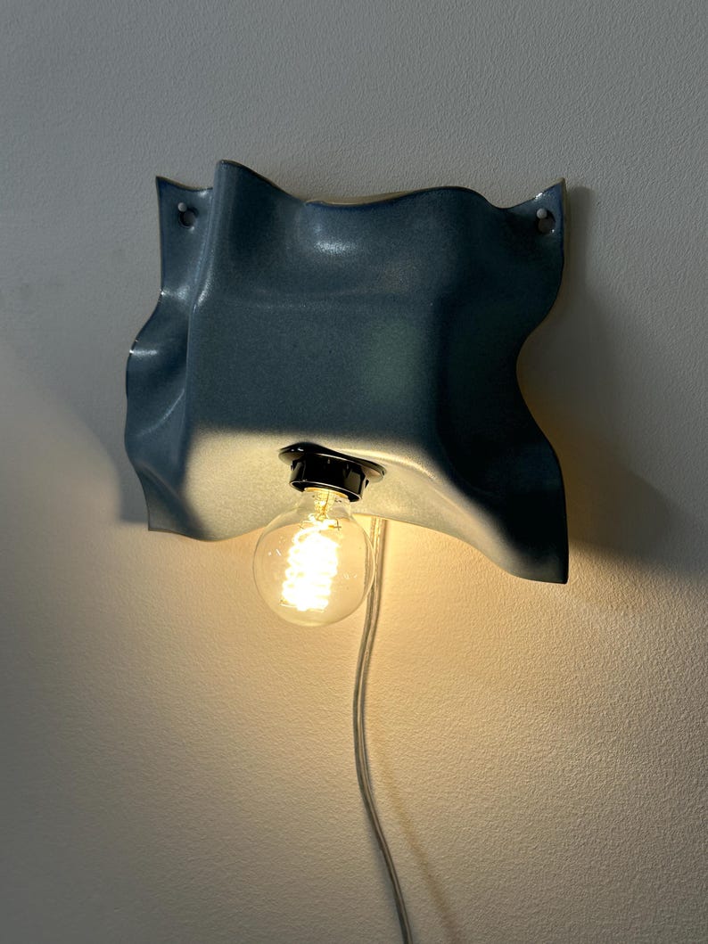 May include: A wall-mounted lamp featuring a blue, wavy, ceramic-like shade with a glossy finish. The fixture includes a black socket holding a glowing, round, clear glass light bulb. A white electrical cord hangs down from the base of the light.