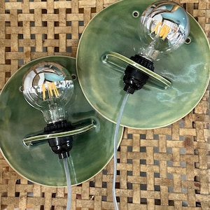 May include: Two wall sconces featuring circular, sage green glazed ceramic backplates. Each fixture includes a black socket holding a clear glass globe light bulb with visible internal filaments and a silver-toned reflective top section.