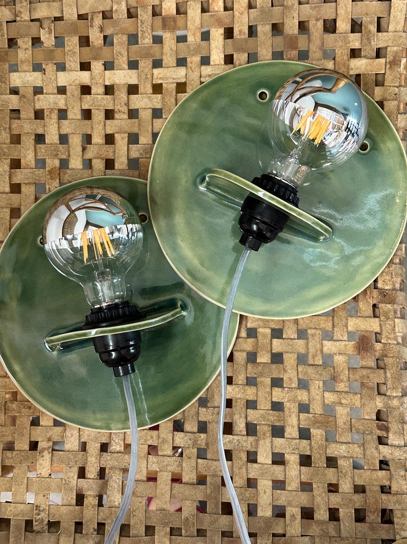 May include: Two wall sconces featuring circular, sage green glazed ceramic backplates. Each fixture includes a black socket holding a clear glass globe light bulb with visible internal filaments and a silver-toned reflective top section.