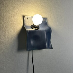 May include: A wall-mounted lamp featuring a curved, dark blue metal sheet with a smooth, glossy finish. A single round, glowing white light bulb is centered in the upper portion of the metal plate, which includes two small holes for mounting.