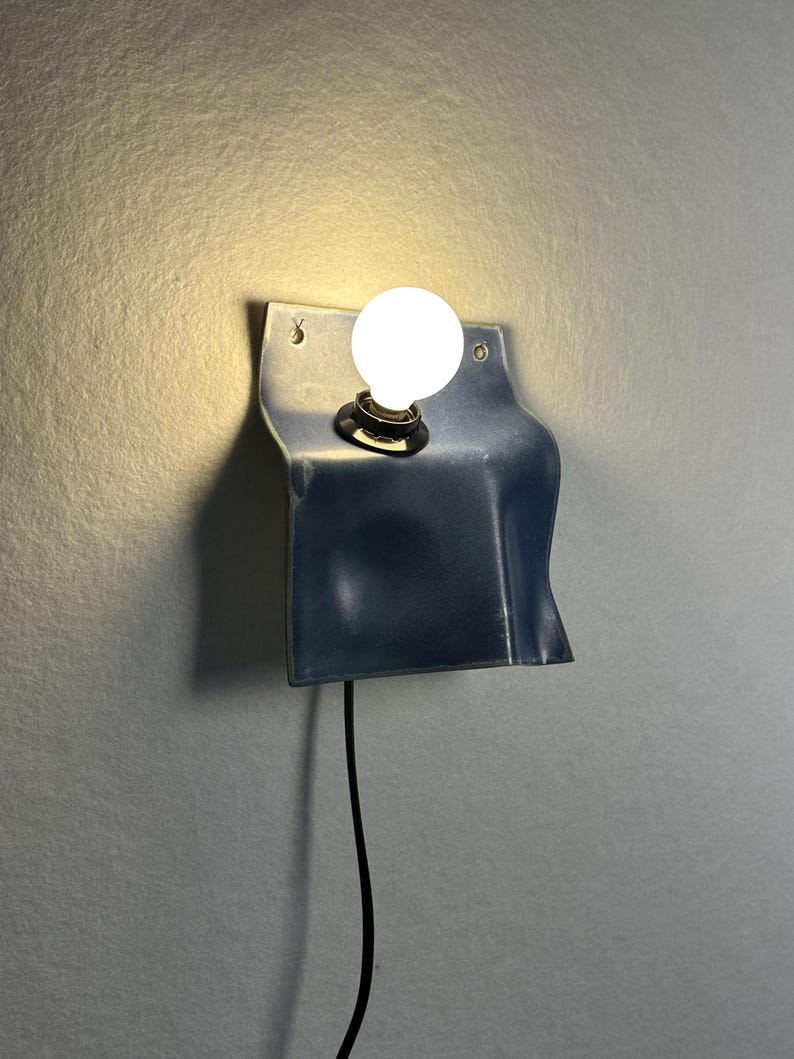 May include: A wall-mounted lamp featuring a curved, dark blue metal sheet with a smooth, glossy finish. A single round, glowing white light bulb is centered in the upper portion of the metal plate, which includes two small holes for mounting.