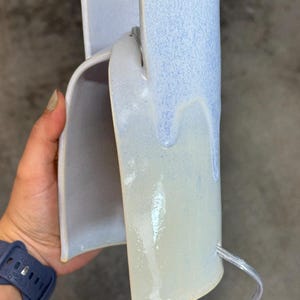 K&ouml;nnte beinhalten: Eine geschwungene Wandleuchte aus Keramik mit einer gl&auml;nzenden Glasur, bei der ein hellblauer oberer Bereich in eine cremefarbene Basis &uuml;bergeht. Die Leuchte verf&uuml;gt &uuml;ber ein transparentes Stromkabel, das unten herausgef&uuml;hrt ist.