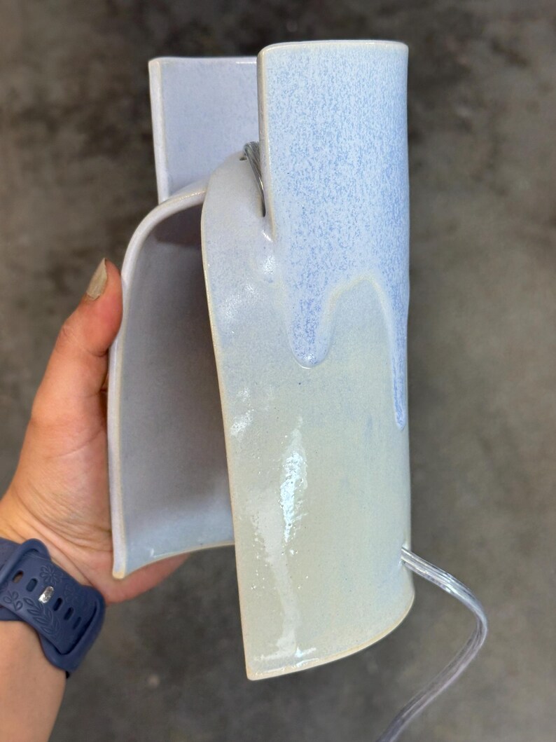 K&ouml;nnte beinhalten: Eine geschwungene Wandleuchte aus Keramik mit einer gl&auml;nzenden Glasur, bei der ein hellblauer oberer Bereich in eine cremefarbene Basis &uuml;bergeht. Die Leuchte verf&uuml;gt &uuml;ber ein transparentes Stromkabel, das unten herausgef&uuml;hrt ist.
