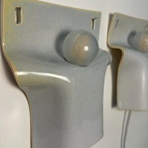May include: Two wall-mounted ceramic light fixtures featuring a smooth, pale blue-grey glazed finish with subtle yellow-tinted edges. Each lamp has a curved, rectangular body with two rectangular cutouts at the top and a single round glass bulb.